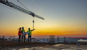 Three Contractors Stand Atop A Building At Dawn, Framed By A Large Crane, Pointing And “looking Toward Tomorrow.”